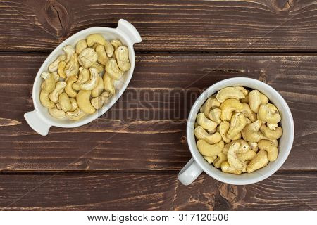 Lot Of Whole Unsalted Beige Cashew In White Oval Ceramic Bowl Flatlay On Brown Wood