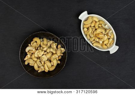 Lot Of Whole Unsalted Beige Cashew In White Oval Ceramic Bowl In Dark Ceramic Bowl Flatlay On Grey S