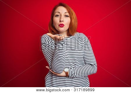 Young redhead woman wearing strapes navy shirt standing over red isolated background looking at the camera blowing a kiss with hand on air being lovely and sexy. Love expression.