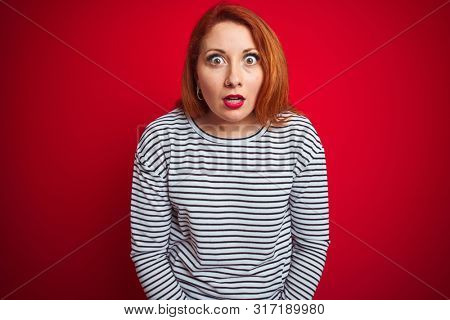 Young redhead woman wearing strapes navy shirt standing over red isolated background afraid and shocked with surprise expression, fear and excited face.