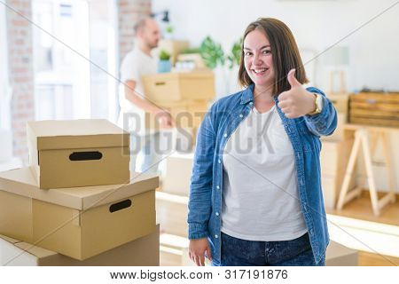 Young couple arround cardboard boxes moving to a new house, plus size woman standing at home doing happy thumbs up gesture with hand. Approving expression looking at the camera with showing success.
