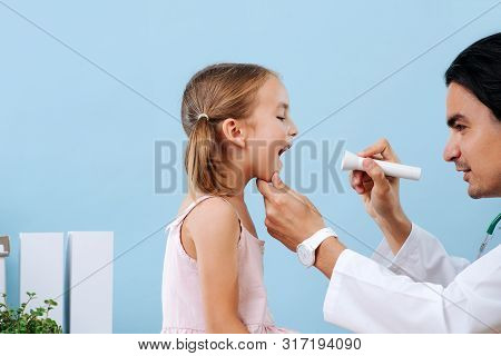 Male Doctor Examining Little Girls Tonsils At Hospital. He Is Using Flashlight, Holding Her Chin. Sh