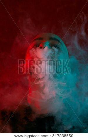 Close-up Studio Shot Of A Young Bald Guy Vaping, Blowing Out A Cloud Of Smoke On Red Background.