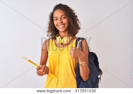 Brazilian student woman wearing backpack holding notebook over isolated white background happy with big smile doing ok sign, thumb up with fingers, excellent sign