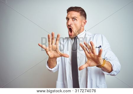 Young handsome doctor man wearing white profressional coat over isolated background afraid and terrified with fear expression stop gesture with hands, shouting in shock. Panic concept.