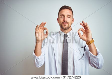 Young handsome doctor man wearing white profressional coat over isolated background relax and smiling with eyes closed doing meditation gesture with fingers. Yoga concept.