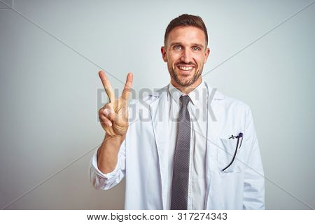 Young handsome doctor man wearing white profressional coat over isolated background showing and pointing up with fingers number two while smiling confident and happy.