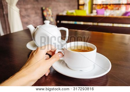 White Cup Of Tea And Teapot On A Table In A Cafe. Female Hand Holds A Cup. The Concept Of Tea Drinki