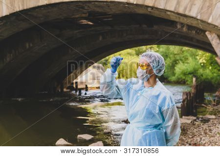 Female Ecologist Or Epidemiologist Takes Test-tube Water Analysis In A City River Under An Old Bridg