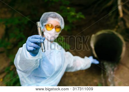 Female Ecologist Or Epidemiologist In A Protective Gown And Mask Demonstrates A Test Tube With A Sam