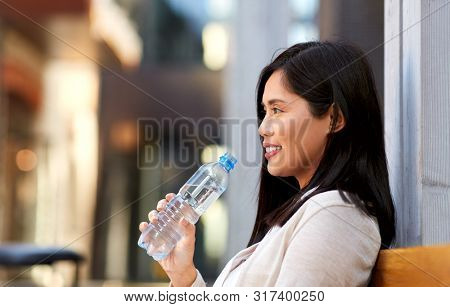 lifestyle and people concept - happy smiling young asian woman drinking water flom plastic bottle sitting on wooden city bench