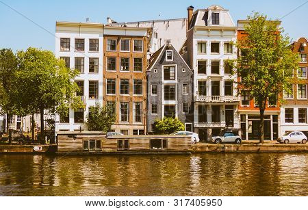 A Houseboat In Front Of A Row Of Pretty Houses Along The Canals In Amsterdam, Netherlands.