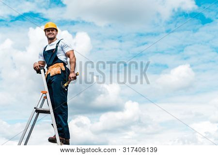 Cheerful Repairman Standing On Ladder And Smiling Against Blue Sky With Clouds