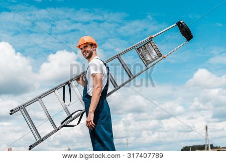 Positive Repairman Holding Ladder And Smiling Against Blue Sky With Clouds