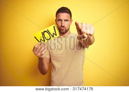 Young man holding wow text banner over yellow isolated background pointing with finger to the camera and to you, hand sign, positive and confident gesture from the front