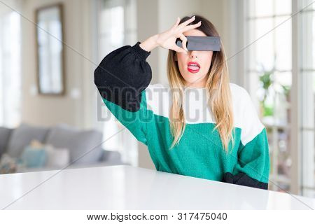 Young beautiful woman blindfold eyes with black censor band at home doing ok gesture with hand smiling, eye looking through fingers with happy face.