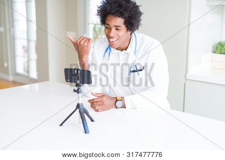 African American doctor man working and talking doing video conferece with smartphone pointing and showing with thumb up to the side with happy face smiling