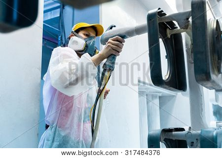Powder Coating Of Metal Parts. A Woman In A Protective Suit Sprays White Powder Paint From A Gun On 