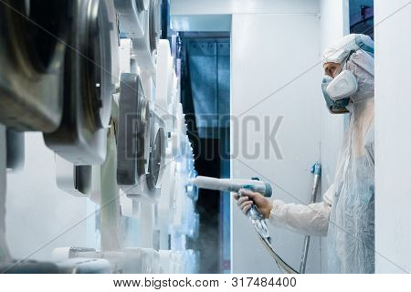 Powder Coating Of Metal Parts. Man In A Protective Suit Sprays White Powder Paint From A Gun On Meta
