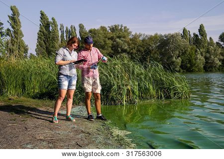 Ecologists On River Bank Examine Sample Of Green Algae And Enter Info On Tablet