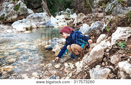A Child With A Backpack Stands Near A Mountain River. The Boy Looks At The Creek. The Kid Walks Thro