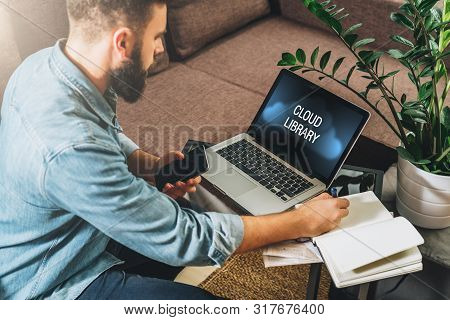 Young Bearded Man Sits At Table, Holds Smartphone, Uses Laptop With Inscription On Monitor- Cloud Li