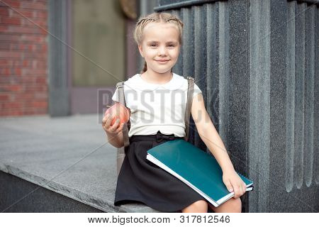 Happy Little Schoolgirl With Blond Hair In School Uniform With Lunch, Book Sits Near School