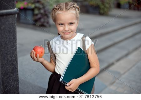 Happy Little Schoolgirl With Blond Hair In School Uniform With Lunch, Book Sits Near School.
