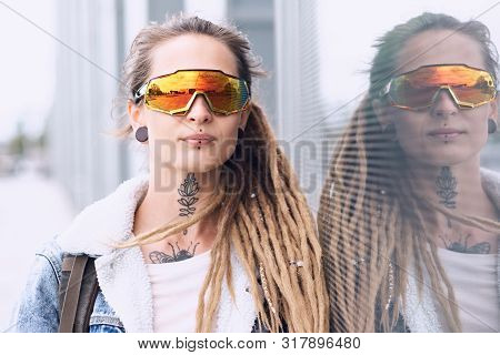 Beautiful Styilish Girl In Big Toned Glasses, Tattoo On The Neck And Piercing Is Posing Near Mirroed