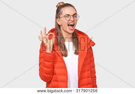 Portrait Of Cheerful Young Woman Wearing Red Jacket, Transparetn Eyeglasses, Showing Ok Gestures Wit