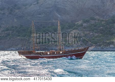 Old Two-masted Yacht In Stormy Sea Against Rocky Shore.
