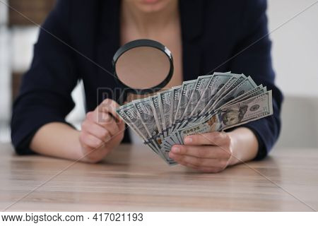 Expert Authenticating 100 Dollar Banknotes With Magnifying Glass At Table In Office, Closeup. Fake M