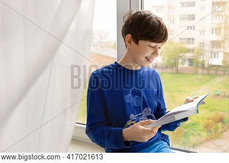 The Child Is Reading A Book. Cheerful Boy Reads A Book At Home On The Windowsill In Rainy Weather