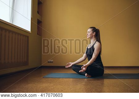 Attractive Woman In Black Sportswear, Practicing Yoga, Is Engaged In Meditation In The Lotus Positio
