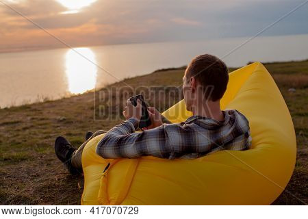 Young Man Sitting On Inflatable Sofa Lamzac With Smartphone While Resting At Sunset On The Mountain
