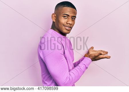Young black man wearing casual pink sweater pointing aside with hands open palms showing copy space, presenting advertisement smiling excited happy 