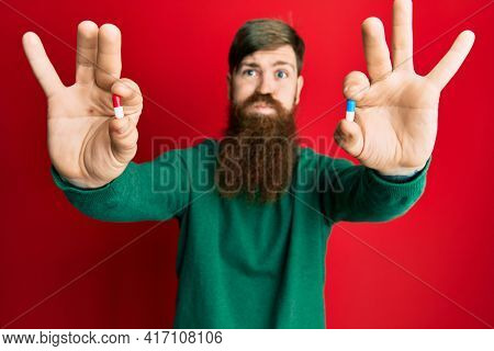 Redhead man with long beard holding two different pills puffing cheeks with funny face. mouth inflated with air, catching air. 