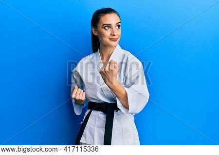 Beautiful brunette young woman wearing karate fighter uniform with black belt doing attack pose smiling looking to the side and staring away thinking. 