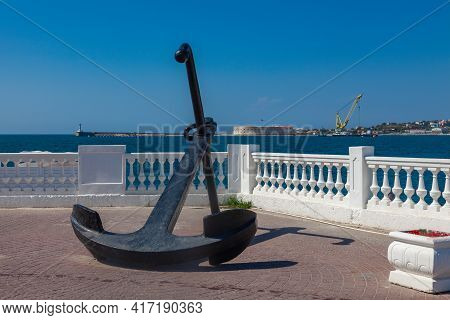 A Large Karable Anchor Lies On The Embankment Against The Background Of A White-stone Fence And The 