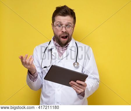 Young Doctor With A Stethoscope Around Neck Holds A Tablet In Hands And Looks At It In Bewilderment.