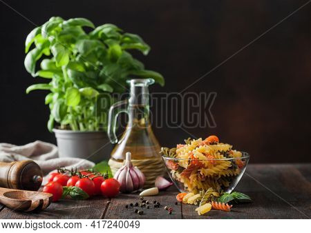 Raw Tricolor Fusilli Pasta In Glass Bowl With Oil And Garlic, Basil Plant And Tomatoes On Wooden Bac