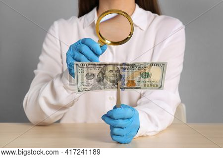 Expert Authenticating 100 Dollar Banknote With Magnifying Glass At Table On Light Grey Background, C