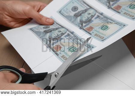 Counterfeiter Cutting Dollar Banknotes With Scissors At Wooden Table, Closeup. Fake Money Concept