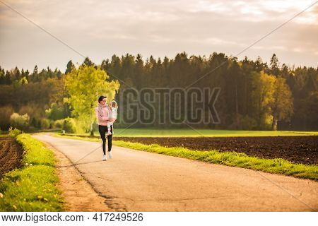 Mother And Child Walking On Countryside Road Between Agricultural Fields Towards Vilage From Forest.