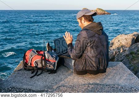 Young Nomadic Traveler Using His Laptop To Make A Video Call With His Family In Front Of The Sea. Bu