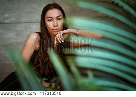 Beautiful Young Woman Closeup Portrait Through Green Leaves Of A Flower On A Gray Wall Background
