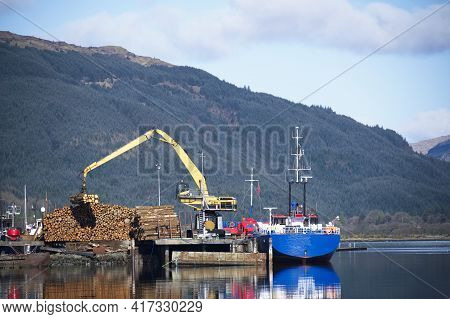 Crane Lifting Wood Logs From Stack To Ship In Sea Loch For Transportation Of Biomass Fuel In Scotlan