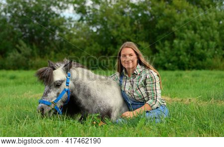 Happy Young Woman Is Sitting On The Grass And Hugging Her Horse.