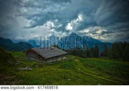 Cloudy And Stormy Day On Alpine Hut On Pasture In Front Of Wood And Mountains With Clouds