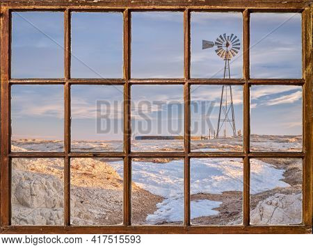 windmill with a pump and cattle water tank in a prairie, winter or early spring scenery as seen from a vintage sash window